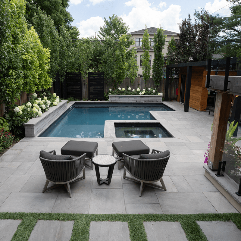 A modern backyard features a rectangular pool, patio area with two chairs, table, and green landscaping surrounded by trees and a wooden structure.
